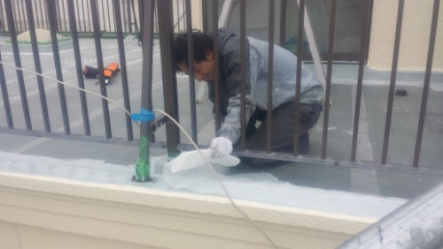 koushin_20140319-1612221 | 外壁塗装は神奈川県横浜市の塗装職人 Maintenance worker kneels on a balcony ledge behind bars, cleaning with a spray bottle and hose.