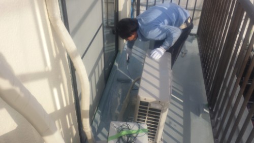 koushin_20140322-1556481 | 外壁塗装は神奈川県横浜市の塗装職人 Maintenance worker on a narrow balcony repairing an outdoor air conditioning unit, wearing gloves and using a tool.
