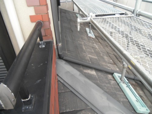koushin_CIMG1153-1 | 外壁塗装は神奈川県横浜市の塗装職人 Scaffolding and a bent handrail on a building exterior with a loose metal panel resting on a sloped roof near a brick wall.