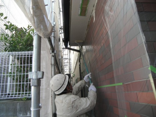 koushin_CIMG1233-1 | 外壁塗装は神奈川県横浜市の塗装職人 Construction worker in a hard hat and gloves applies green tape to the exterior brick wall during renovation, with scaffolding visible nearby.