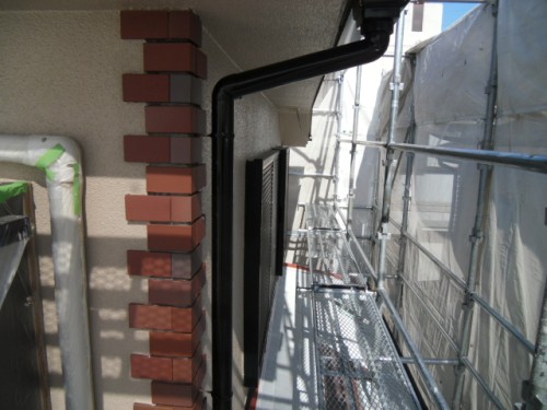 koushin_CIMG1240-1 | 外壁塗装は神奈川県横浜市の塗装職人 Construction site along a beige building: scaffolding extends along the wall on the right, with a brick column on the left and a black downspout visible between them.