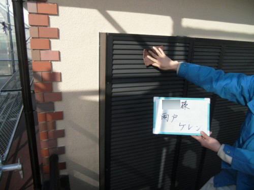 koushin_CIMG1244-1 | 外壁塗装は神奈川県横浜市の塗装職人 Person in a blue jacket reaches to open a mailbox slot on a black metal surface, holding a white card with Japanese writing, outdoors beside a brick corner.