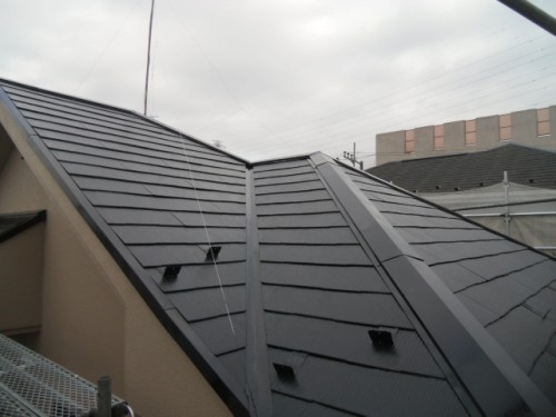 koushin_CIMG1248-1 | 外壁塗装は神奈川県横浜市の塗装職人 Close-up view of a dark metal-tiled roof with ridge satisfying joints; scaffolding and a building under construction visible in the background.