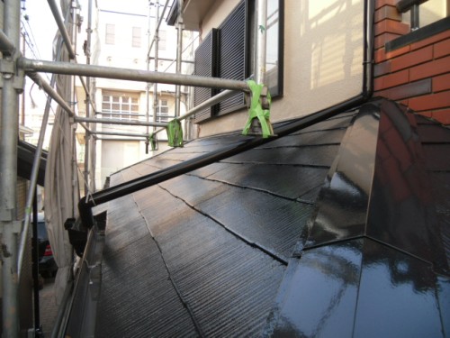 koushin_CIMG1263-1 | 外壁塗装は神奈川県横浜市の塗装職人 Scaffolding runs along a sloped black tiled roof with green clamps and nearby buildings.