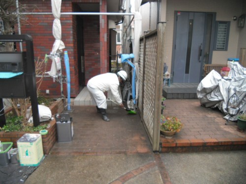 koushin_CIMG1265-1 | 外壁塗装は神奈川県横浜市の塗装職人 Worker in white protective suit and helmet kneels to repair ground near a gate in a narrow residential alley; construction supplies and scaffolding nearby.