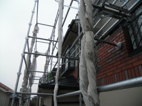 koushin_CIMG1266-1 | 外壁塗装は神奈川県横浜市の塗装職人 Scaffolding surrounding a brick house with wrapped poles and metal frames on the exterior.