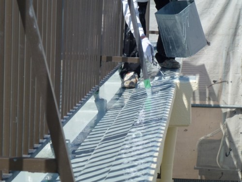 koushin_P10208251 | 外壁塗装は神奈川県横浜市の塗装職人 Worker wearing gloves paints a metal railing along a sunlit roof edge; a paint bucket and tray sit nearby.