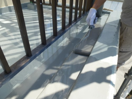 koushin_P10208281 | 外壁塗装は神奈川県横浜市の塗装職人 Worker applying gray waterproof sealant along a rooftop edge with a roller, beside a metal railing.