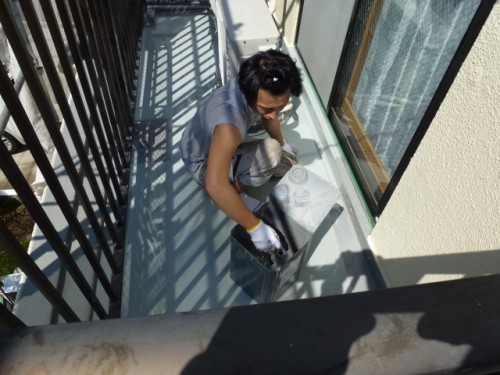 Person kneeling on a narrow balcony, painting the floor with a roller and tray beside the sliding door.