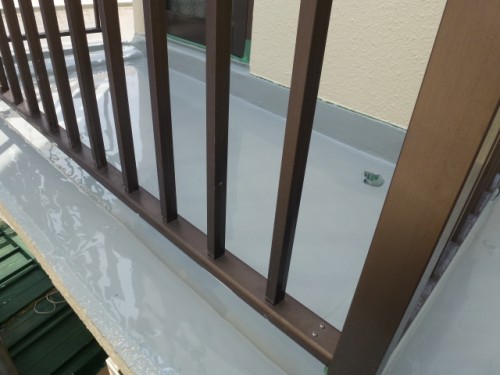 koushin_P10208391 | 外壁塗装は神奈川県横浜市の塗装職人 Close-up of a brown metal balcony railing along a white waterproof edge beside a beige exterior wall.