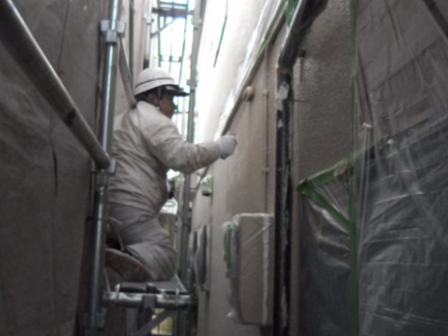 koushin_gaiheki_uwanuri | 外壁塗装は神奈川県横浜市の塗装職人 Construction worker in a hard hat and protective clothing on scaffold applying material to a building wall in a narrow alleyway.