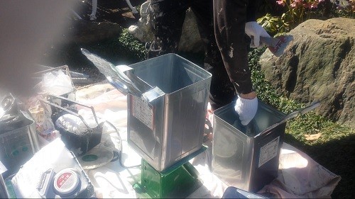 koushin_keiryou1 | 外壁塗装は神奈川県横浜市の塗装職人 Outdoor workshop scene with a person wearing gloves placing items into a metal bin mounted on a small green platform, surrounded by tools and bags.
