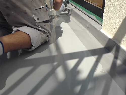 koushin_top-hake | 外壁塗装は神奈川県横浜市の塗装職人 Construction worker kneeling on a gray rooftop surface, applying sealant with a tool near a door; shadowed railing crosses the area.