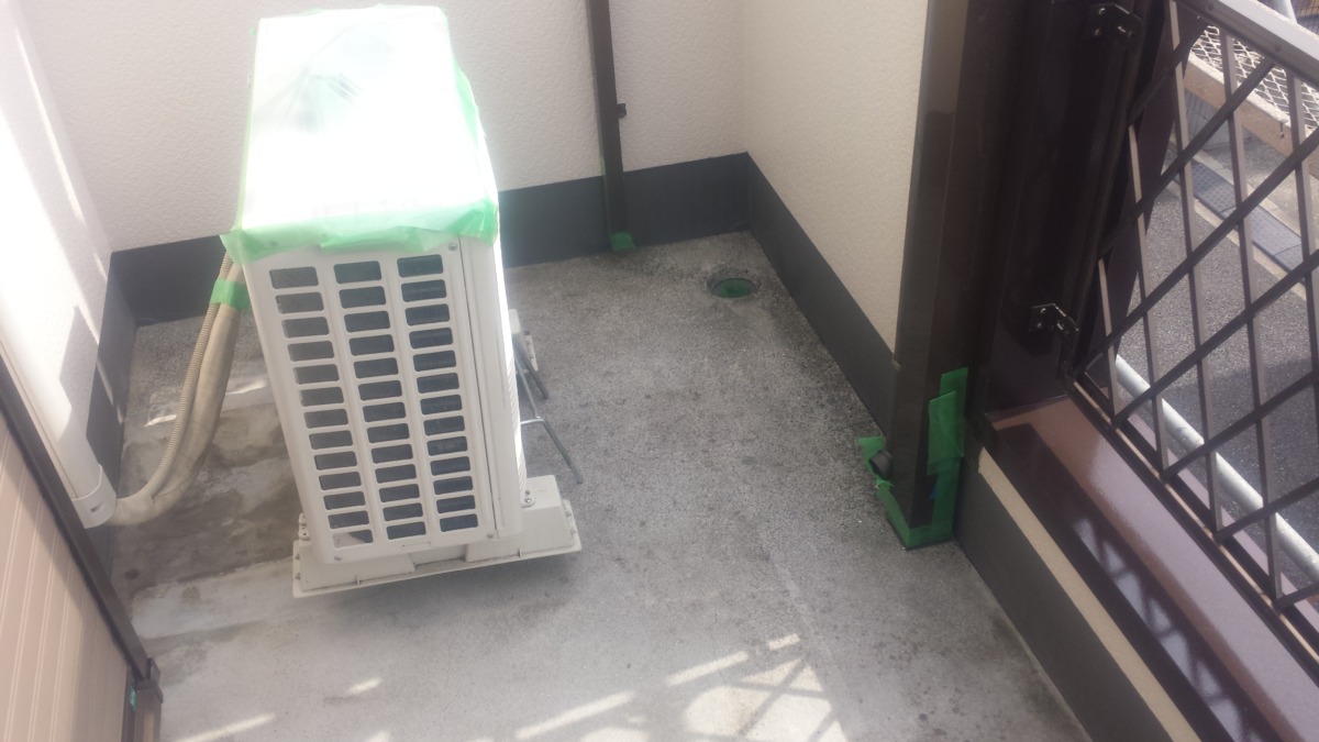 METADATA-START | 外壁塗装は神奈川県横浜市の塗装職人 Outdoor air conditioner/condenser unit on a small balcony, partially wrapped in green protective cover, with a drain hole and railing nearby.