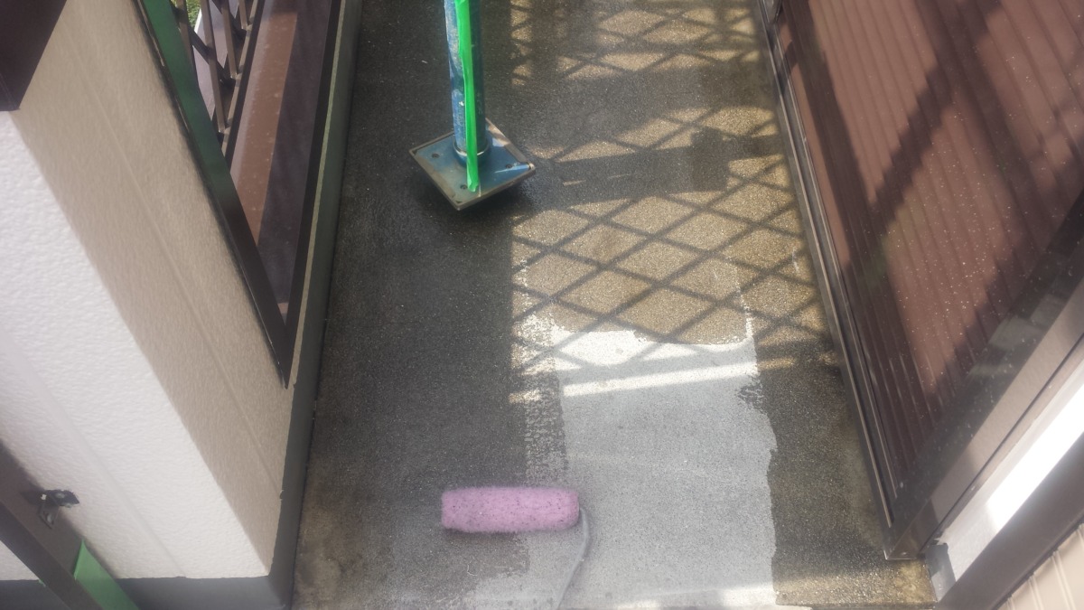 METADATA-START | 外壁塗装は神奈川県横浜市の塗装職人 Narrow balcony with a pink paint roller on a wet gray concrete floor; a green pole and diamond-shaped railing shadows nearby.