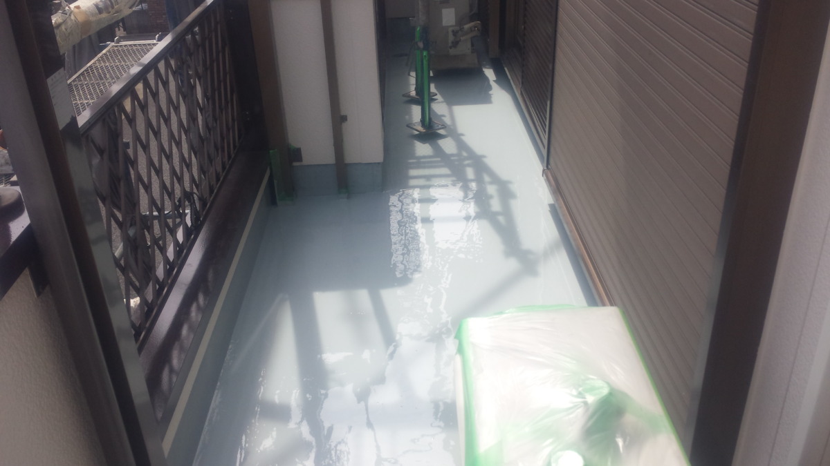 METADATA-START | 外壁塗装は神奈川県横浜市の塗装職人 Narrow outdoor balcony with a wet light-gray epoxy floor and plastic-covered item in the foreground, construction scaffolding shadows visible.