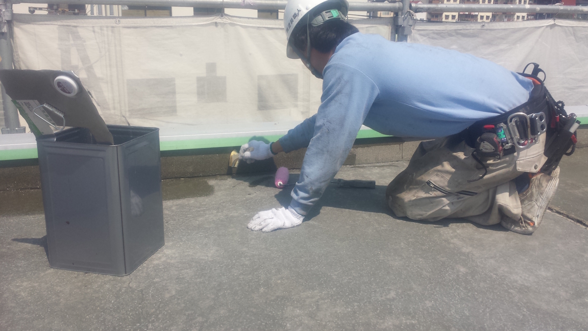 METADATA-START | 外壁塗装は神奈川県横浜市の塗装職人 Construction worker kneeling on a roof, wearing a helmet and gloves, using a small polishing tool near a trash can and railing edge.