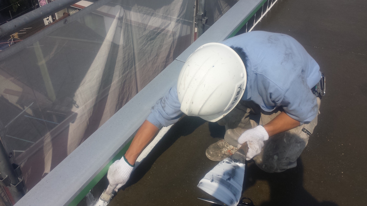 METADATA-START | 外壁塗装は神奈川県横浜市の塗装職人 Construction worker in a white helmet and blue shirt applies sealant along a railing on a flat rooftop edge.
