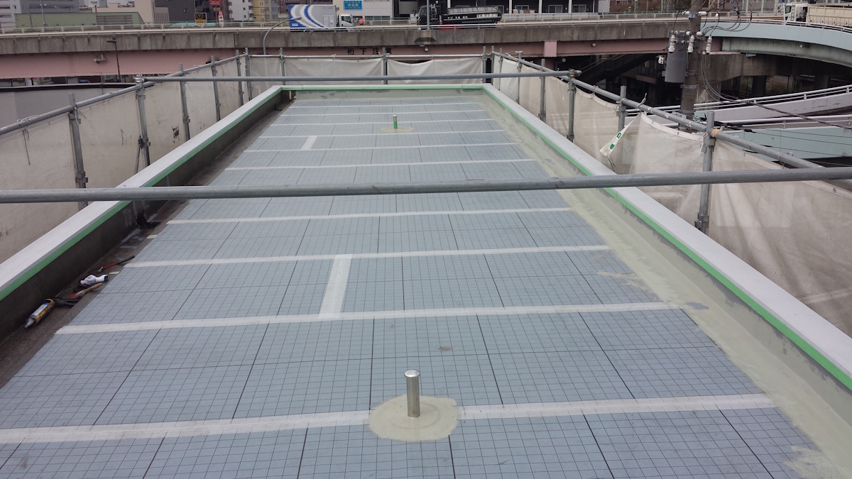 METADATA-START | 外壁塗装は神奈川県横浜市の塗装職人 Rooftop under construction with blue grid tiles, scaffolding along the edges, and a single vent in the foreground; city buildings and a highway overpass in the distance.