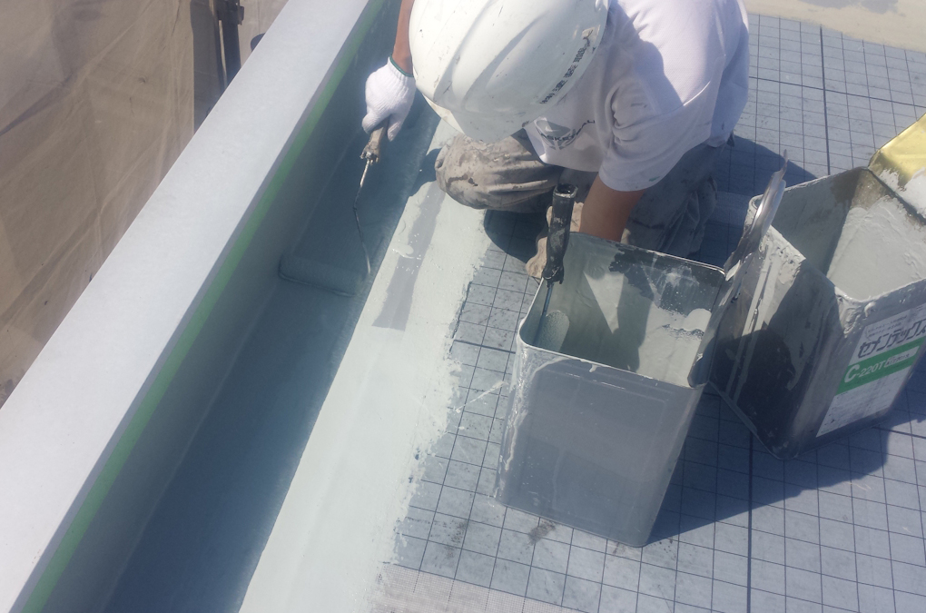 METADATA-START | 外壁塗装は神奈川県横浜市の塗装職人 Construction worker in a white helmet applies light gray waterproof coating along a roof edge using a roller, with buckets nearby.