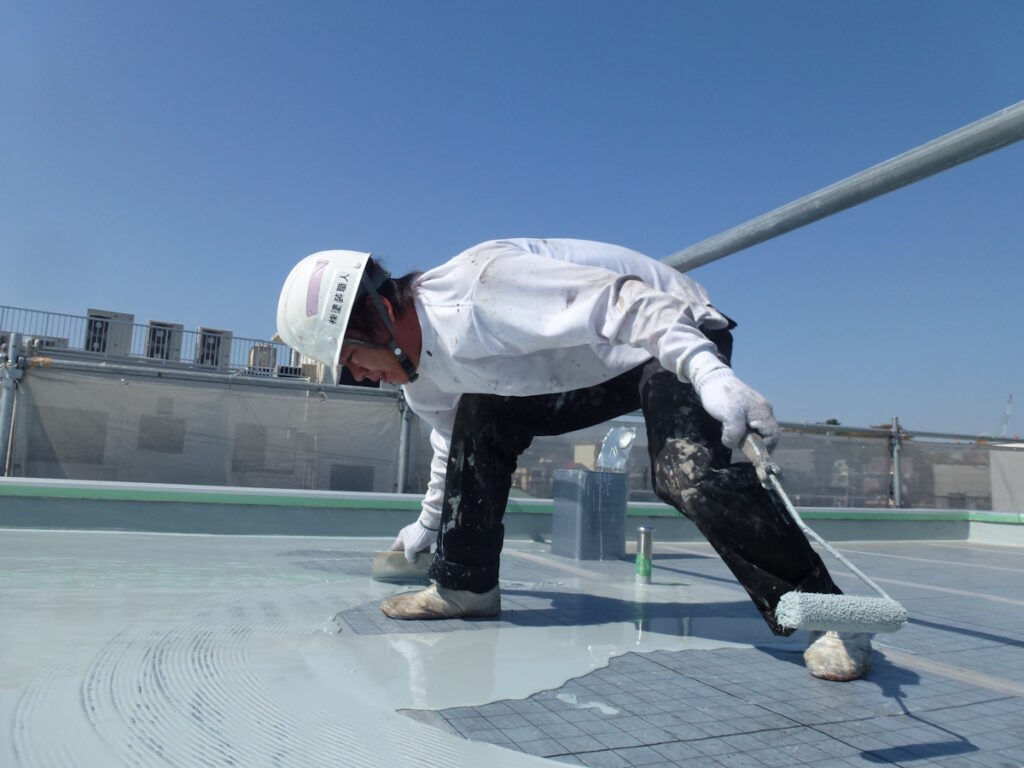 P1030226 | 外壁塗装は神奈川県横浜市の塗装職人 Construction worker wearing a white helmet and gloves rolls a protective roof coating across a flat rooftop on a bright day.