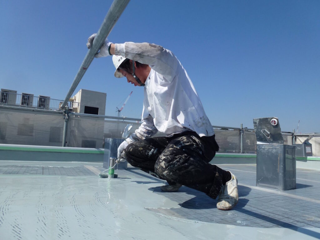 P1030234 | 外壁塗装は神奈川県横浜市の塗装職人 Worker kneels on a rooftop, applying a liquid coating with a long roller under a clear blue sky, wearing a helmet and gloves.