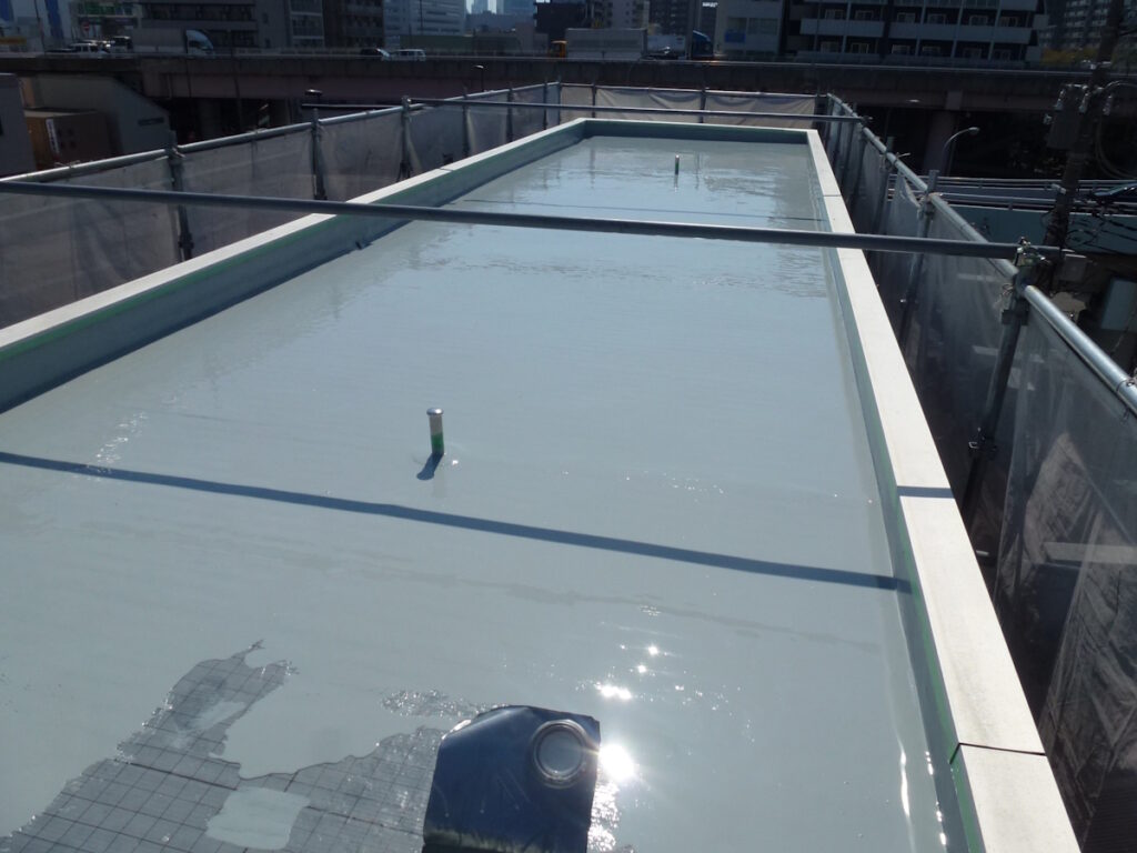 P1030239 | 外壁塗装は神奈川県横浜市の塗装職人 Rooftop deck with a shallow pool of water on a pale blue surface, framed by scaffolding and city buildings beyond.