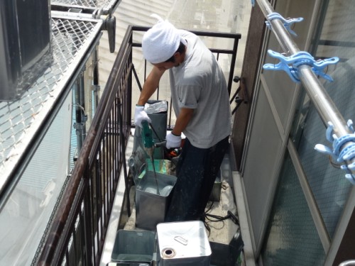 P1030274-1 | 外壁塗装は神奈川県横浜市の塗装職人 Construction worker on a narrow balcony using a handheld angle grinder to cut metal near the railing, wearing gloves and a white head wrap
