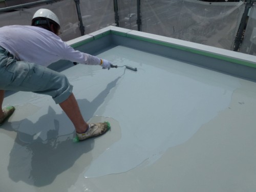 P1030281-1 | 外壁塗装は神奈川県横浜市の塗装職人 Worker wearing a helmet and gloves applying a gray liquid waterproof coating to a flat rooftop using a trowel/roller