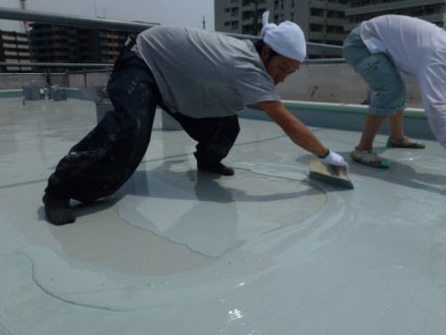 P1030298-1 | 外壁塗装は神奈川県横浜市の塗装職人 Two workers kneel and spread a light-colored liquid coating on a flat roof, tools in hand, wearing head coverings and gloves.