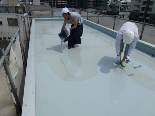 P1030307-1 | 外壁塗装は神奈川県横浜市の塗装職人 Two workers on a rooftop coating a flat roof with liquid sealant, one pouring from a can while the other smooths with a roller, both wearing hard hats.