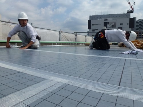 koushin_P1030141-1 | 外壁塗装は神奈川県横浜市の塗装職人 Two construction workers in hard hats kneel on a tiled rooftop under construction, with a building and crane in the background.