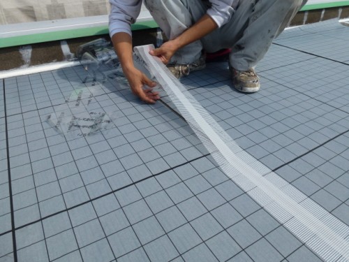 koushin_P1030150-1 | 外壁塗装は神奈川県横浜市の塗装職人 Worker kneels on a grid-patterned surface, laying and taping a seam in underlayment.