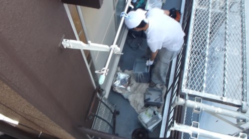 koushin_netaba-1 | 外壁塗装は神奈川県横浜市の塗装職人 Maintenance worker in a white shirt and cap on a narrow balcony, handling tools and inspecting a toolbox.