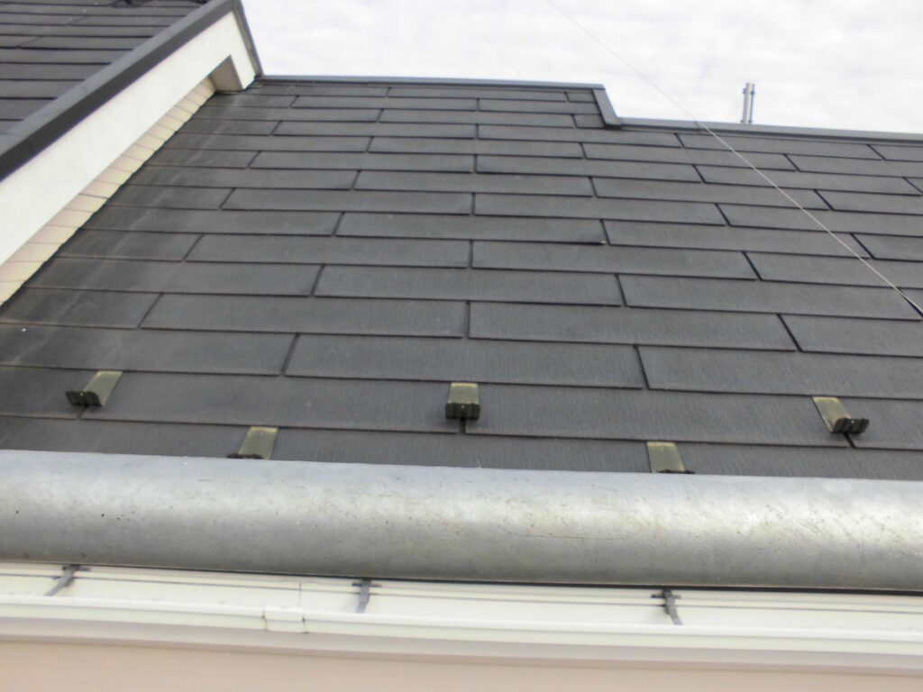 Dark slate roof with rectangular shingles, seen from below, with a metal gutter pipe in the foreground and white fascia at the bottom.