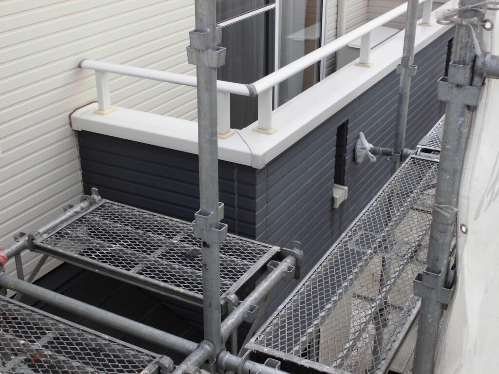 Construction scaffolding along a house exterior with metal platforms and guard rails, showing light-colored siding and a dark blue/gray wall paneling detail.