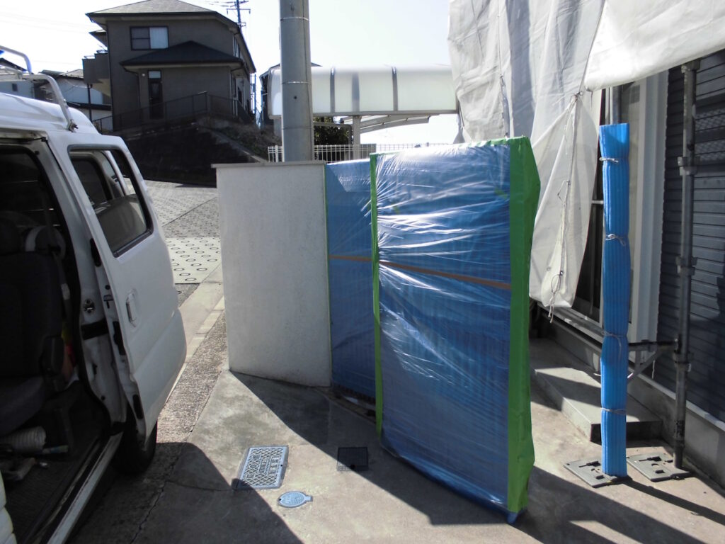 Van with open sliding door parked on a sidewalk beside a blue plastic‑wrapped construction panel secured with green tape
