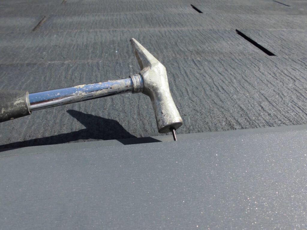 Close-up of a hammer with a blue handle resting on a gray roof, a nail protruding from the edge near the head.