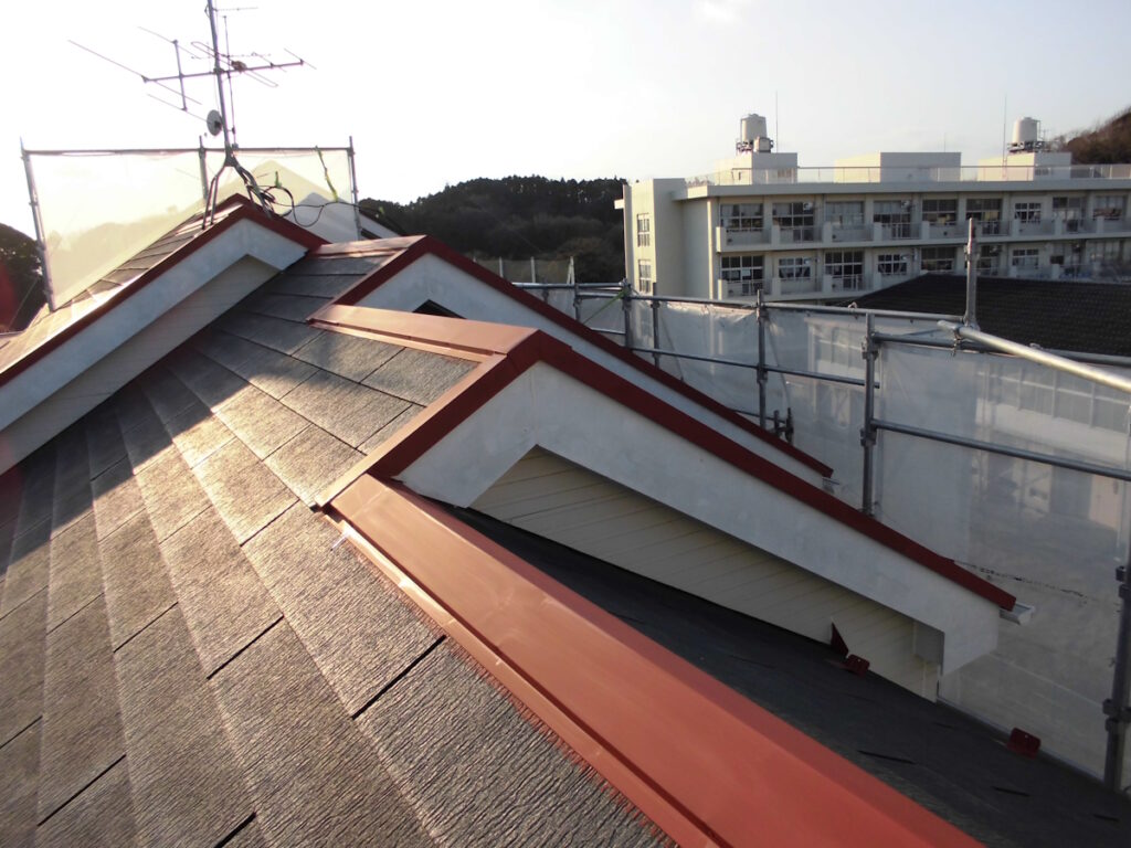 Sloped rooftop with gray shingles and red metal trim, scaffolding along the edge, with a white building in the distance.
