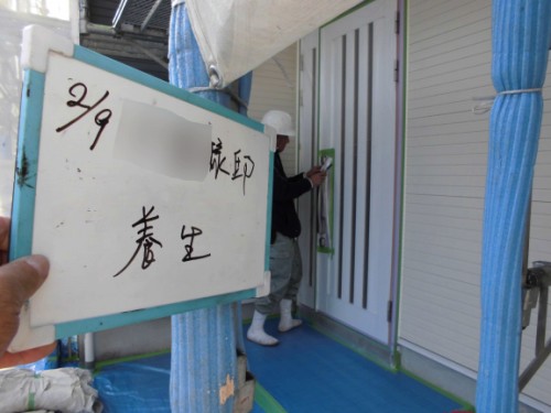 Construction workers install a white door at a building under construction; a hand holds a sign with Japanese writing and a date in front of the doorway.