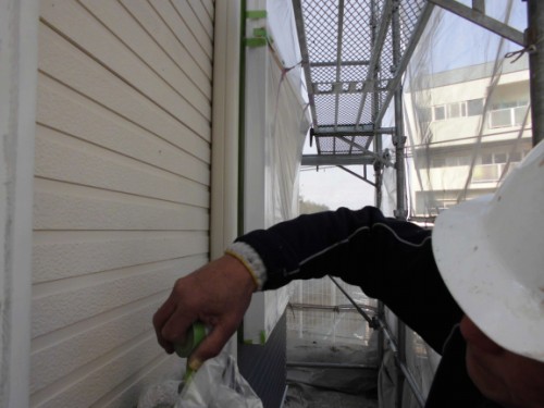 Construction worker in a white hard hat on scaffolding, applying sealant to the building exterior with a green-handled tool.