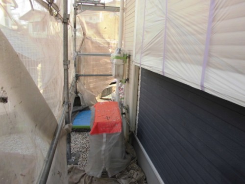 Narrow construction site between buildings with scaffolding and plastic sheeting; a red-covered rectangular object sits on a platform beside scattered materials.