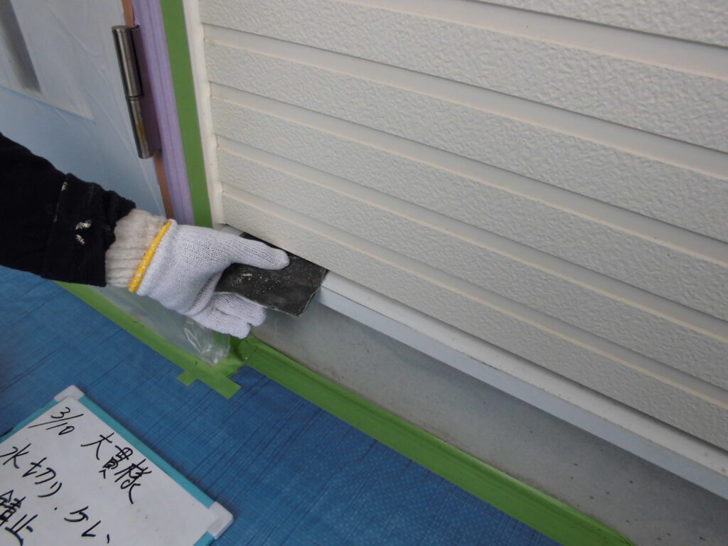 Gloved hand presses a black sealant strip along the bottom edge of white siding.