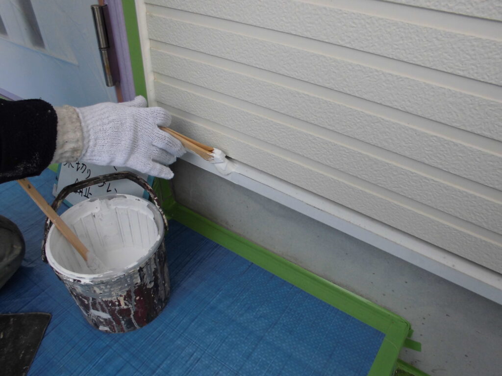 Gloved hand smoothing sealant along the base of vinyl siding with a putty knife; a paint bucket and blue tarp are nearby.