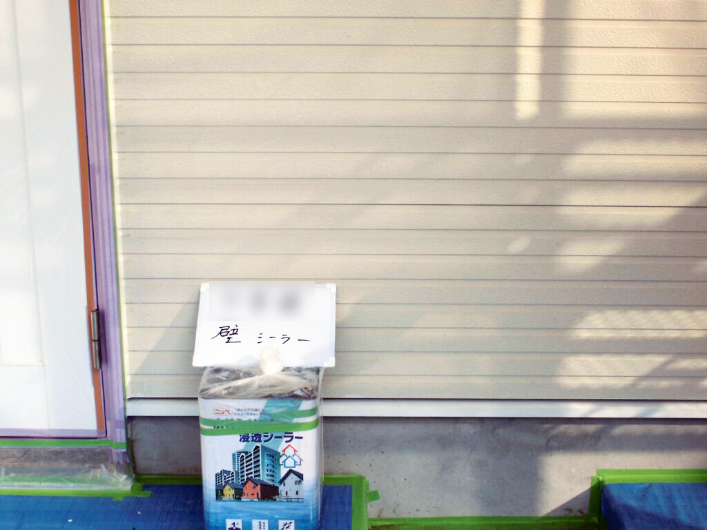 Bag of sealer with a white handwritten sign on top, placed in front of a closed beige garage door; sign has Japanese writing and the bag shows a cityscape graphic.