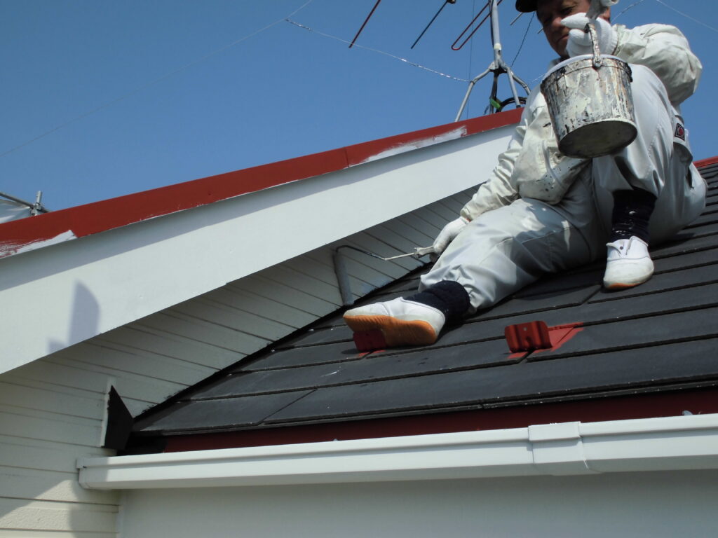 Worker in white coveralls sits on a sloped roof, painting the white trim with a paint bucket nearby against a blue sky.