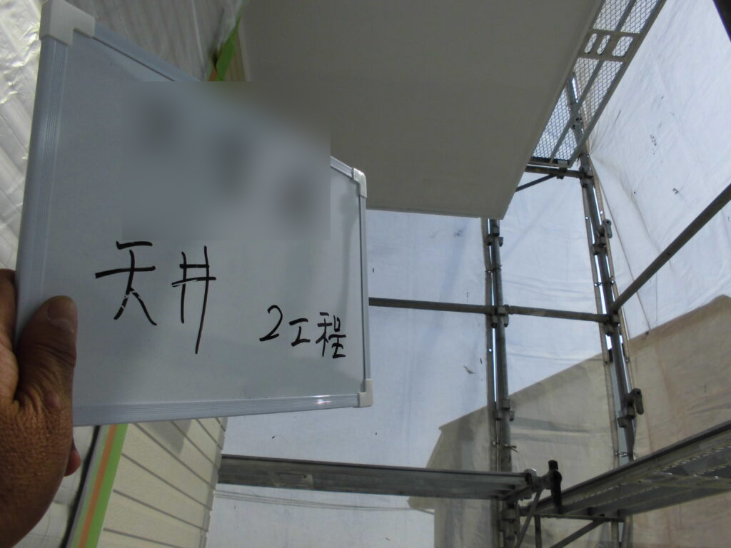 Hand holding a whiteboard with handwritten Japanese text at a construction site with metal scaffolding in the background.