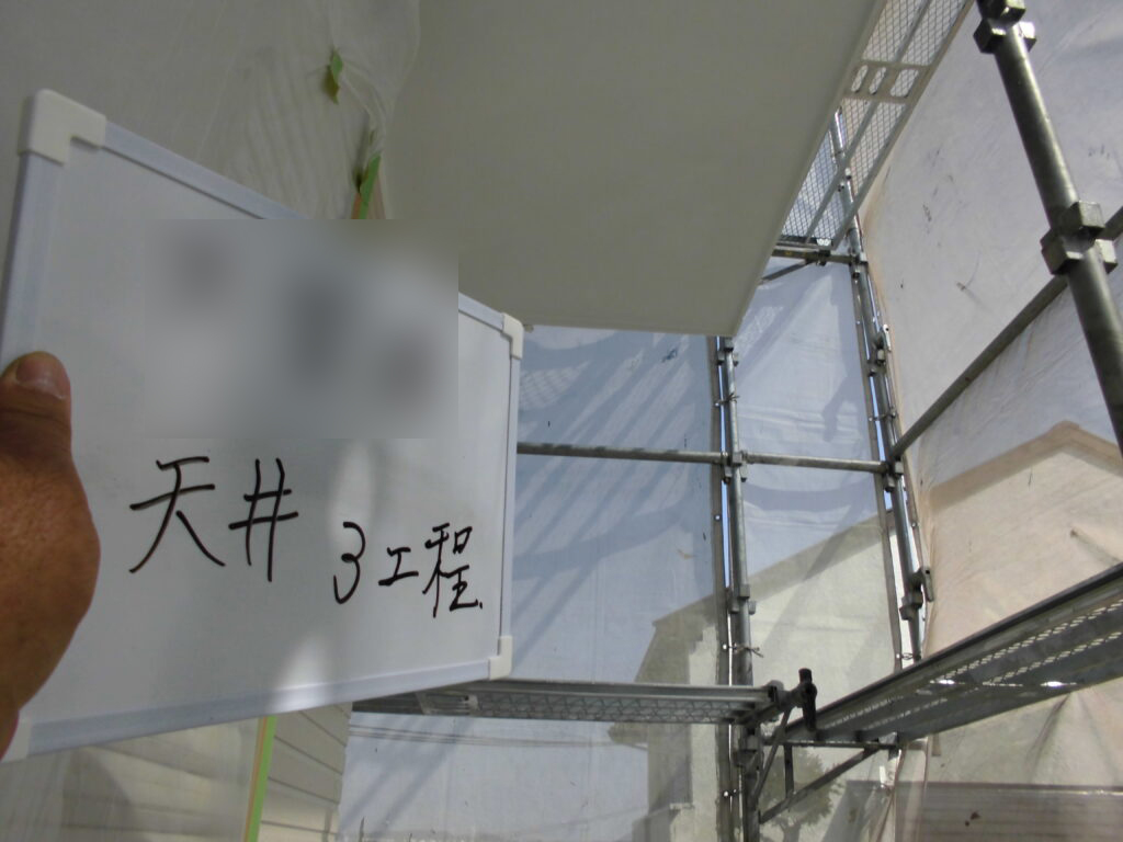 Hand holding a whiteboard with blurred top area and handwritten Chinese/Japanese characters (天井 3工程) in front of a metal scaffolding construction site backdrop.