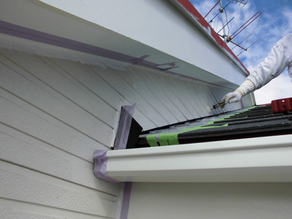 Worker wearing a glove applying sealant along the white gutter edge beside light siding, with masking tape and green-sashed shingles nearby.