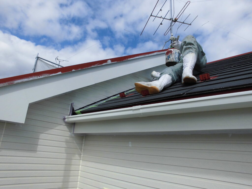Worker in protective clothing sits on a sloped roof, painting the edge with a paint can in hand on a sunny day.